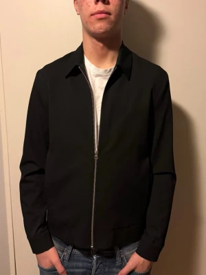 Svart bomberjacka från Les Deux - Nypris 2799! Svart bomberjacka från Les Deux med stilren design och silverfärgad dragkedja framtill. Jackan har klassisk krage, raka ärmar och diskret logga på bröstet. Materialet är mjukt och lätt, perfekt för dig som gillar minimalistisk stil.