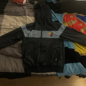 Svart vindjacka Djurgården - Svart och ljusblå vindjacka med huva från Djurgården. Jackan har klubbens emblem på bröstet, hel dragkedja framtill och två fickor med dragkedja. Perfekt för dig som vill visa ditt lagstöd och hålla dig skyddad mot vinden. Jackan köptes för 6 år sedan limiterad så de finns ingen som har den:)buda nått