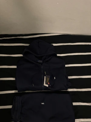 Mörkblå mjukisbyxor och hoodie från Polo Ralph Lauren - Säljer ett par mörkblå mjukisbyxor från Polo Ralph Lauren med klassisk vit logobrodyr på benet. Byxorna har ribbade muddar och är gjorda i mjukt bomullsmaterial, perfekta för chill dagar. Snygg och enkel design med skön passform. Hör av dig om du har några frågor