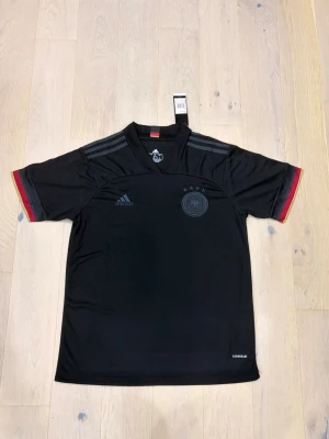 Svart Adidas fotbollströja Tyskland - Säljer en svart Adidas fotbollströja med Tysklands landslagsmärke och fyra stjärnor på bröstet. Tröjan har grå ränder på axlarna och detaljer i rött och gult vid ärmsluten. Materialet är lätt polyester och perfekt för fotboll.