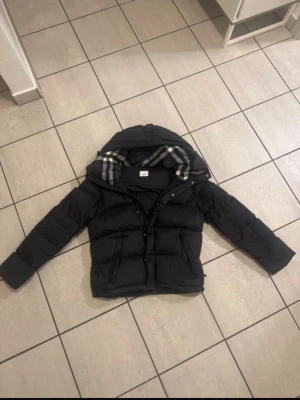 Svart pufferjacka från Burberry - Svart pufferjacka från Burberry med avtagbar huva och klassiskt rutigt foder. Jackan har stora vita logopatchar på ärmen och insidan, dragkedja och knappar framtill samt två fickor med dragkedja. Perfekt för kalla dagar och riktigt snygg streetstil.