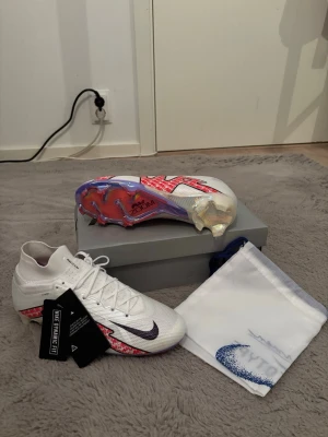Nike Mercurial Vapor Elite FG fotbollsskor - Säljer ett par Nike Mercurial fotbollsskor med Vaporposite+ och ACC-teknologi. Skorna är vita med röda och svarta detaljer, har snörning och en platt sula med iriserande undersida. Materialet är syntet och mesh, och mönstret är randigt. Perfekt för dig som vill ha grym touch på planen.