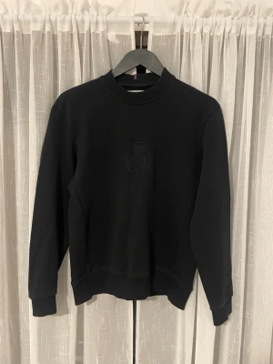 Svart sweatshirt från Les Deux - Svart sweatshirt från Les Deux med diskret broderad logga på bröstet. Tröjan har rund hals, ribbade muddar och är långärmad. Materialet är mjuk bomull som ger en skön känsla. Perfekt för dig som gillar stilrena och enkla plagg.