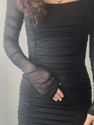 Svart långklänning med meshärmar - Säljer en svart långklänning med transparenta meshärmar och snygg rynkad design längs hela klänningen. Klänningen har en figurnära passform och lång ärm, vilket ger en cool och modern look. Perfekt för dig som gillar stilrena och trendiga plagg.