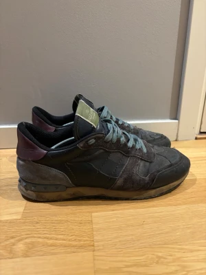 Valentino Garavani Rockrunner sneakers - Säljer ett par Valentino  Rockrunner sneakers i mörka toner med svarta och grå paneler, vinröd häl och gröna skosnören. Skorna har detaljerade nitar på sulan och är tillverkade i mocka och skinn. Snygg och unik design som sticker ut.
