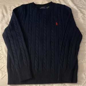 Mörkblå kabelstickad tröja Polo Ralph Lauren - Kabelstickad mörkblå tröja från Polo Ralph Lauren med klassisk röd broderad logga på bröstet. Tröjan har rund halsringning, långa ärmar och ribbade muddar. Perfekt för dig som gillar tidlös och stilren design.