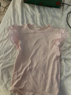 Ljusrosa topp med volangärmar - Säljer en ljusrosa topp med ribbad struktur och korta ärmar med romantiska volangdetaljer. Ärmarna har broderade hålmönster som ger en söt och feminin touch. Perfekt för dig som gillar mjuka färger och detaljer.