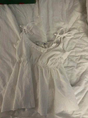 Vit linnetopp med knytband - Säljer en söt vit linnetopp från 157 med knytband på axlarna och volangkant vid halsen. Toppen har en luftig passform och är tillverkad i ett lätt, strukturerat material som ger en fräsch känsla. Perfekt för varma dagar och enkel att matcha.