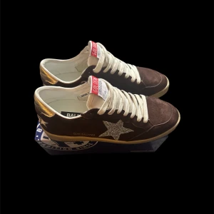 Golden Goose Ball Stars - Snygga Golden Goose Ball Star sneakers i mörkbrunt mocka med vit snörning och glittrig stjärndetalj på sidan. Hälen är metallic guld och sulan är beige. Tungan har rödvit etikett med logga. Perfekt för dig som gillar unika detaljer och cool stil.