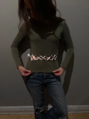 Occasio - Säljer en olivgrön långärmad topp med croppad passform och snygg snörningsdetalj i vitt band framtill midjan. Toppen har en tight siluett och är tillverkad i ett stretchigt material som sitter skönt på kroppen. Perfekt för dig som vill sticka ut med en unik detalj. (Tokyo) 🌟storlek S