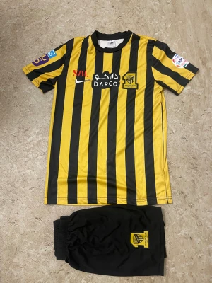 Randig fotbollströja Benzema #9 - Säljer en gul och svart randig fotbollströja från Nike med ITTIHAD CLUB-logga och Benzema #9 på ryggen. Tröjan har flera sponsorer tryckta på bröstet och ärmarna. Tillhörande svarta shorts med klubbmärke ingår. Perfekt för dig som älskar fotboll och vill ha en snygg matchtröja.