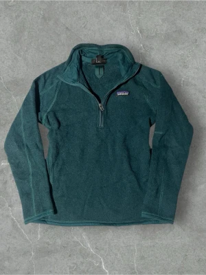 Mörkgrön fleece-tröja Patagonia | 36/S - Mysig mörkgrön fleece-tröja från Patagonia med halv dragkedja framtill och diskret logga på bröstet. Tröjan har lång ärm, hög krage och en praktisk ficka med dragkedja på sidan. Perfekt för kyliga dagar och outdoor-häng.