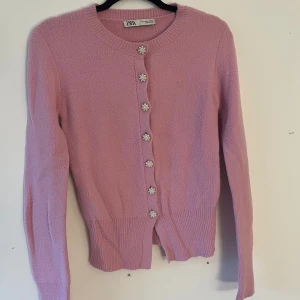 Rosa kofta från Zara med pärlknappar - Superfin ljusrosa kofta från Zara med lång ärm och rund hals. Snygga blomformade pärlknappar framtill ger en gullig touch. Mjuk stickad känsla och ribbad nederkant. Perfekt för dig som gillar söta detaljer och vill ha något som sticker ut.