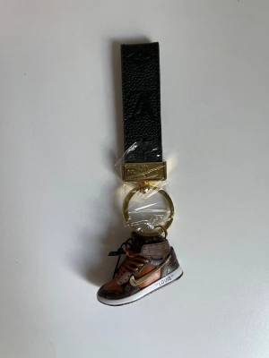Nyckelring med Vuitton sneaker - Snygg nyckelring med svart läderrem och guldfärgad metallring. Hängande detalj i form av en miniatyrsneaker i brunt med klassiskt Vuitton-mönster och orange snörning. Perfekt accessoar för dig som gillar streetstyle och lyxiga detaljer.