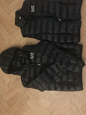Svart jacka och dunväst EA7 Emporio Armani - Säljer en svart jacka och dunväst från EA7 Emporio Armani. Jackan och västen har quiltad design, dragkedja framtill och en stilren logga på bröstet. Materialet är syntet och dun, vilket gör den lätt och varm. Perfekt för dig som vill ha en clean look med lite extra edge.