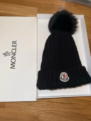 Svart mössa med pälsboll - Snygg svart  mössa med stor fluffig pälsboll på toppen och ribbstickad design. Använts fåtal gånger men super fin. Kan tänkas gå ner i pris vid snabb affär.