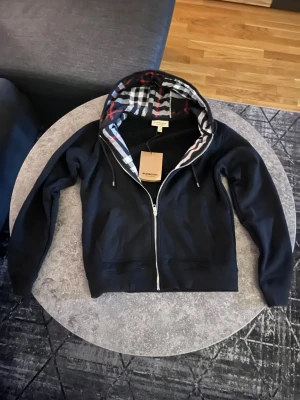 Svart Burberry Hoodie - Svart hoodie från Burberry i storlek M, tillverkad i 100% bomull. Huva med klassiskt Burberry-rutigt mönster i svart, vit, grå och röd. Dragkedja framtill, snörning vid huvan och två fickor. Perfekt för dig som gillar stilrena streetwear-vibbar.