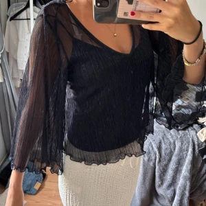 Mörkblå glittrig blus med vida ärmar - Supersnygg svart blus med glittriga detaljer och transparenta, vida ärmar. Blusen har v-ringning och volangkant nertill och vid ärmslut. Perfekt för dig som vill sticka ut lite extra med en elegant och trendig look.