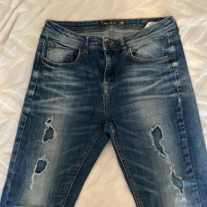 Blå slitna jeans från Amisu - Säljer ett par blå jeans från Misu med snygga slitningar och hål på båda benen. Jeansen har klassisk femficksdesign, normal midja och raka ben. 