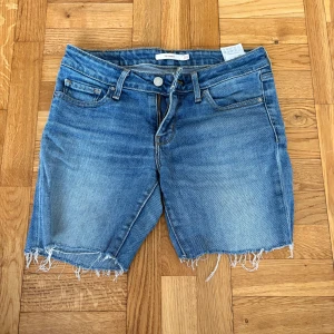Levi's jeansshorts med rå kant - Snygga blå jeansshorts från Levi's, modell 711 Skinny, med klassisk femficksdesign och rå, fransig kant nedtill. Shortsen har normal passform och midwaist, tillverkade i mjuk bomull med dragkedja och knapp framtill. Perfekta för en chill sommardag.