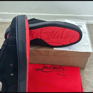 Svarta sneakers från Christian Louboutin - Säljer ett par svarta sneakers från Christian Louboutin med ikonisk röd sula och snygg signatur på undersidan. Skorna har rund tå, platt sula och snörning. Ovandelen är i mjuk mocka och insidan är beige. Perfekt för dig som gillar exklusiv streetstyle.