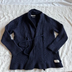 Mörkblå stickad kofta Odd Molly - Mysig mörkblå stickad kofta från Odd Molly med knytband i midjan och diskret mönsterstickning. Koftan har lång ärm och en liten tygpatch nertill. Perfekt för lager-på-lager och chill vibes. Skick: tröjan är en blandning av ull, alpacka och något mer (som inte är läsbart (se etiketten)), tröjan är något användning- och tvättpåvekad (se bilder) . Storlek : storlek 3 står angivet, men jag tycker den är något mindre i storleken, en medium skulle jag säga, typ 38 eller 36.  Mjuka lager och spets.