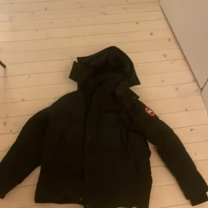 Svart dunjacka från Canada Goose S - Säljer en svart dunjacka från Canada Goose i storlek S. Jackan har huva, klassisk logga på ärmen och stängs med både dragkedja och knappar. Perfekt för kalla dagar och riktigt snygg streetstil.
