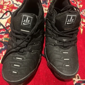 Svarta Nike Air Vapormax Plus - Säljer ett par svarta Nike Air Vapormax Plus sneakers med cool design och tydliga loggor på plösen. Skorna har snörning och en futuristisk look med detaljer i svart och reflekterande inslag. Perfekta för dig som gillar streetstyle och vill ha något unikt på fötterna.