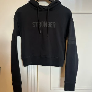 Svart hoodie från Stronger - Svart croppad hoodie från Stronger med tryckt logga på bröstet och snörning i huvan. Långa ärmar med diskreta ränder på ena ärmen. Perfekt för chill eller träning, mjukt material och relaxed fit. Pris kan diskuteras!