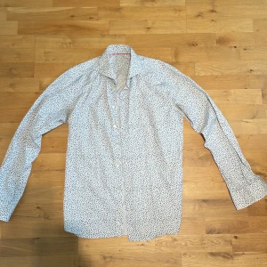 Vit blommig skjorta från Eton - Snygg vit skjorta från Eton i slim fit med små blå och svarta blommor över hela tyget. Skjortan har klassisk krage, långa ärmar och knäppning framtill. Perfekt för dig som vill sticka ut lite extra med ett coolt mönster. (Sitter som en Large).