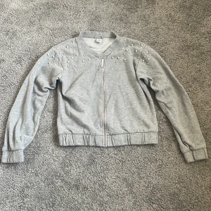 Grå bomberjacka med pärlor från Lindex - Grå bomberjacka från Lindex med dragkedja framtill och pärldetaljer på axlarna. Jackan har ribbade muddar och är tillverkad i mjuk bomullsmix. Perfekt för dig som gillar en chill och snygg stil.