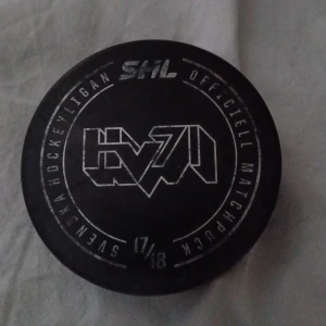SHL Officiell Matchpuck HV71 17/18 - Officiell matchpuck från SHL-säsongen 2017/18 med HV71-logga. Pucken är i gott skick med några ytliga repor, perfekt för samlare eller hockeyentusiaster. En unik souvenir från Svenska Hockeyligan.
