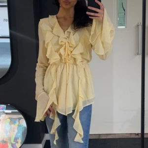 Gul volangblus  - Supersöt ljusgul blus med volanger framtill och längs ärmsluten. Blusen har knytning vid bysten och v-ringning, samt långa, vida ärmar. Tunt och luftigt material som passar perfekt till jeans eller kjol.