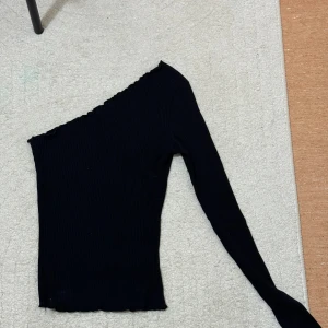 Svart oneshoulder topp från NA-KD - Säljer en svart ribbad oneshoulder topp från NA-KD i storlek XS. Toppen har lång ärm på ena sidan och är av stretchigt material som sitter snyggt på kroppen. Perfekt för dig som gillar en trendig och stilren look.