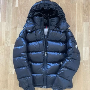 Moncler Maya Svart Jacka  - Säljer en svart pufferjacka från Moncler med glansig finish, vadderad design och avtagbar huva. Jackan har dragkedja framtill, fickor med dragkedja och en ärmficka med Moncler-logga. Perfekt för kalla dagar och riktigt snygg streetstil. Använd 1 vinter med inga defekter eller hål, Storlek 2 som motsvarar M, hör av er vid minsta fundering 🤩🙌