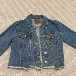 Jeansjacka med nitar från Amisu - Säljer en blå jeansjacka från Amisu med knappar på kragen och bröstfickorna. Jackan har rå kant nertill och klassiska knappar framtill. 