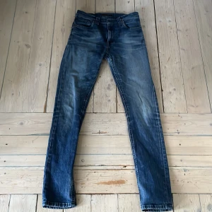 Blå skinny jeans från Nudie Jeans - Säljer ett par blå skinny jeans från Nudie Jeans med klassisk femficksdesign och snygga slitningar. Jeansen är tillverkade i 100% ekologisk bomull och har en mörk tvätt med ljusa partier. Perfekta för dig som gillar en smal passform och stilren look. Storlek w28 L32