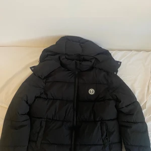 Svart pufferjacka från Trapstar L - Svart pufferjacka från Trapstar med avtagbar huva och dragkedja framtill. Jackan har en clean look med Trapstar-logga på bröstet och stora vadderade paneler. Perfekt för kalla dagar och riktigt streetwear-vibe. Kontakta mig vid behov av fler bilder!