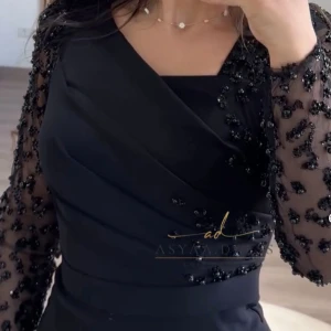 Oanvänd svart långklänning Asyaa Dress - Elegant svart långklänning från Asyaa Dress med figurnära passform. Klänningen har långa transparenta ärmar dekorerade med svarta pärlor och paljetter, samt omlottdetaljer upptill. Perfekt för dig som vill sticka ut med stilrena detaljer och lyxig känsla.
