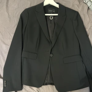 Svart kavaj från STOCKH LM, strl 38 - Snygg svart kavaj från STOCKH LM i klassisk modell med smal passform. Kavajen har en knapp framtill, två fickor med lock och stilren krage. Fodret är svart med diskret logga. Perfekt att styla med sneakers för en cool look. (Byxorna finns också om man vill ha det som komplett)!!