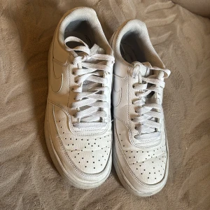 Nike Air Force 1 vita sneakers - Klassiska Nike Air Force 1 sneakers i helvitt skinn med perforerad tå och rund tå. Skorna har snörning och den ikoniska swoosh-loggan på sidorna. Perfekt för dig som gillar en clean och tidlös stil.