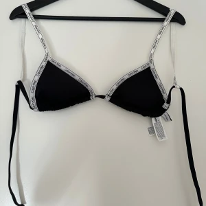 Svart bikiniöverdel från Calvin Klein - Svart bikiniöverdel från Calvin Klein med vita band och logotyp längs kanterna. Triangelmodell med smala axelband och knyt i ryggen. Snygg och stilren design som passar perfekt till stranden eller poolen. Aldrig använd 😍