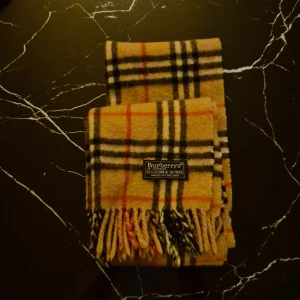 Rutig halsduk från Burberrys i kashmir - Klassisk halsduk från Burberrys i beige, svart, röd och vit rutig design. Gjord av 50% kashmir och 50% ull, med fransar i ändarna. Den ikoniska brittiska stilen gör den till ett statement-plagg för vintern. Knappt använd.