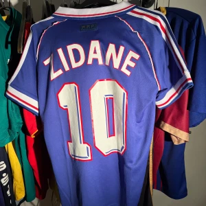 Frankrike Zidane VM 98 fotbollströja - Legendary blå fotbollströja från Frankrike med Zidane och nummer 10 på ryggen. Adidas-logga, FIFA World Cup France 98-tryck och klassiska vita och röda ränder. Broderat landslagsmärke på bröstet. Perfekt för dig som älskar retro och ikoniska spelare. Inte äkta 
