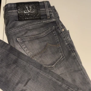 Grå jeans från Jacob Cohën, stl 30 - Snygga grå jeans från Jacob Cohën med klassisk femficksdesign och coolt broderat J på myntfickan. Jeansen har en mörkgrå tvätt och detaljerad svart patch bak i midjan. Perfekt passform och stilren look för dig som gillar premiumjeans.liten defekt som inte märks av alls 