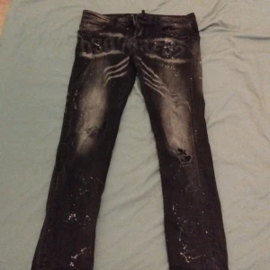 Svarta Dsquared2 jeans med tryck - Säljer ett par svarta jeans från Dsquared2 med coolt tryck och slitningar framtill. Jeansen har vita färgstänk och en skinny passform. Materialet är klassisk denim och de har en unik streetstyle-vibe med detaljerade slitningar och tryck över låren.