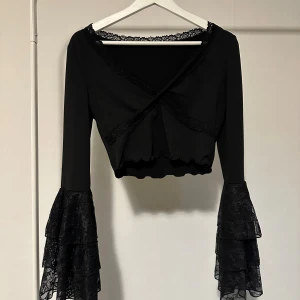 Svart omlott blus med spets och volangärm - Säljer en svart omlott blus med croppad passform och långa ärmar med flera lager volang i spets. Blusen har spetsdetaljer längs halsringningen och omlottfronten, vilket ger en extra cool vibe. Perfekt för dig som gillar statement-ärmar och snygga detaljer.