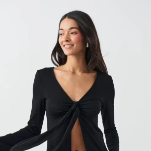 Svart långärmad omlottopp med knyt - Svart långärmad topp från Gina Tricot med djup v-ringning och knyt framtill. Toppens material är mjukt och stretchigt, vilket ger en skön passform. Perfekt för dig som gillar trendiga och stilrena plagg med en twist.