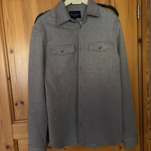 Grå overshirt från Bondelid - Snygg grå overshirt från Bondelid med diskret mönster och klassisk krage. Två bröstfickor med knappar och långa ärmar. Perfekt att slänga över en t-shirt för en chill look. Materialet känns mjukt och har en lite tjockare känsla, vilket gör den extra skön.