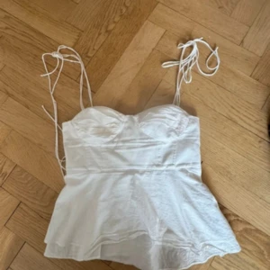 Vit korsettliknande topp med knyt - Säljer en vit korsettinspirerad topp med tunna axelband som knyts upptill. Toppen har markerade kupor och lätt utsvängd nederdel. Baksidan har smockdetaljer för extra komfort. Perfekt för dig som gillar en feminin och trendig look.